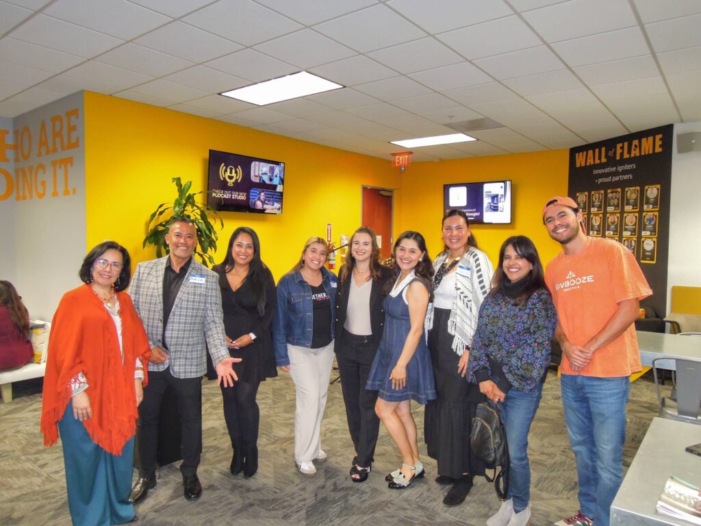 A group of professionals standing together in a modern workspace, smiling and posing in front of a vibrant yellow wall with a "Wall of Flame" display. The group is part of a networking or collaborative event, showcasing diverse business people working together.